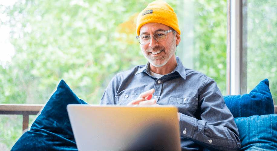Man wearing a yellow beanie and glasses, sitting on a blue couch, using a laptop.