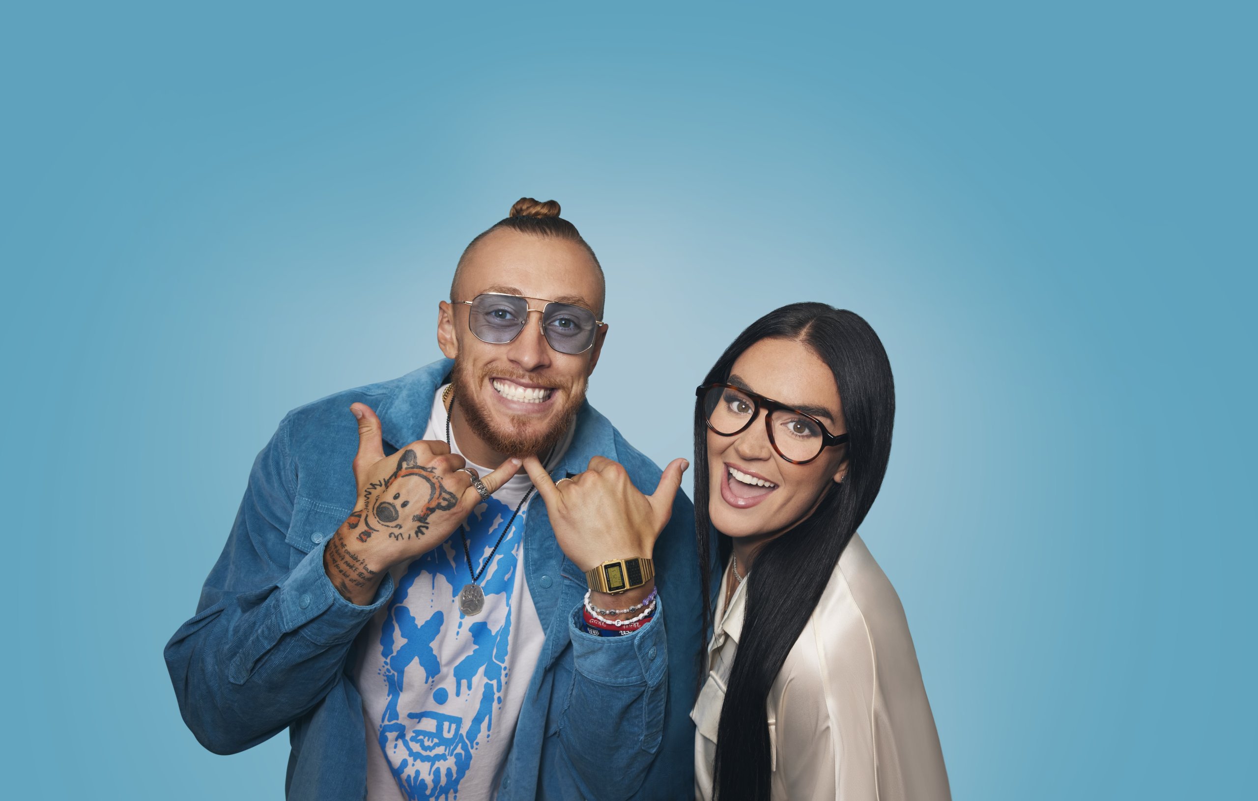 Two smiling people wearing glasses; one has tattoos and a blue jacket, the other with long black hair.