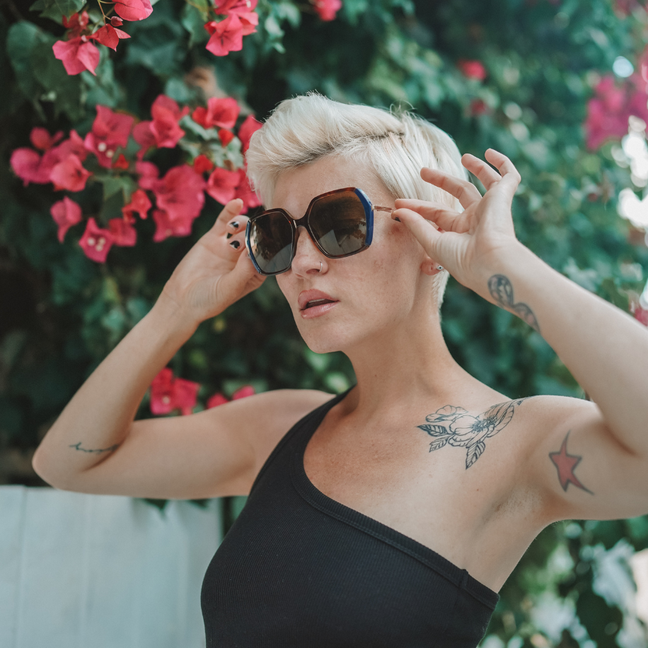 Woman wearing oversized sunglasses with dark lenses, adjusting them with both hands against a floral background.