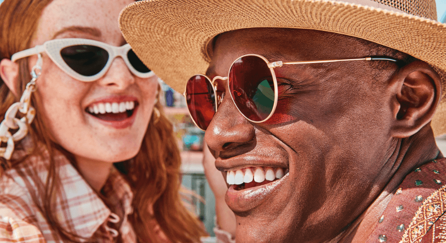 Smiling man wearing round, red-tinted sunglasses and a straw hat, beside woman in white cat-eye sunglasses.