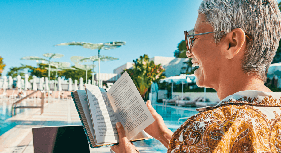 Person with gray hair reading a book near a swimming pool, smiling and wearing glasses.