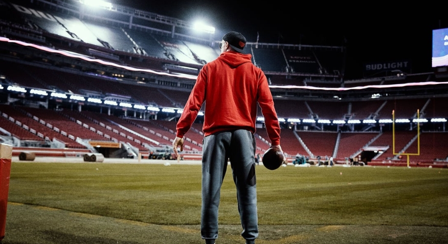 Person wearing a red hoodie and gray sweatpants holding a football on an empty, lit-up football field.