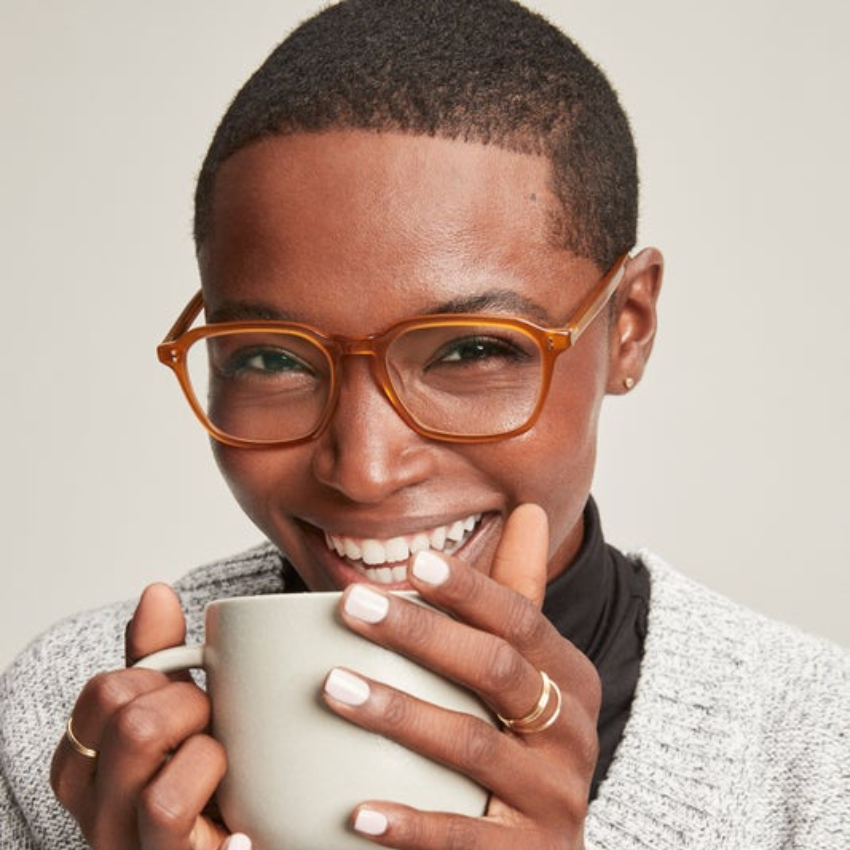 Person wearing orange glasses and holding a gray ceramic mug with both hands while smiling.