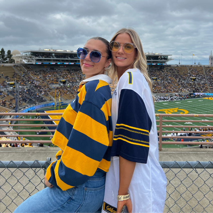 Two women wearing blue and yellow sports jerseys with sunglasses, posing at a football stadium.