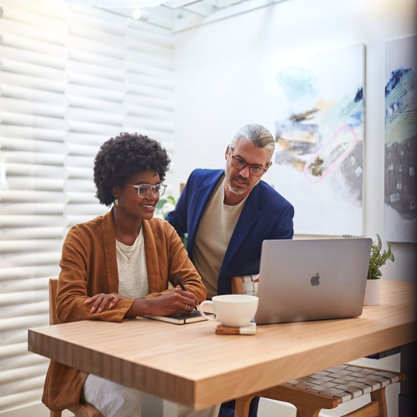 Two people collaborating at a wooden table with an Apple laptop and a white mug.