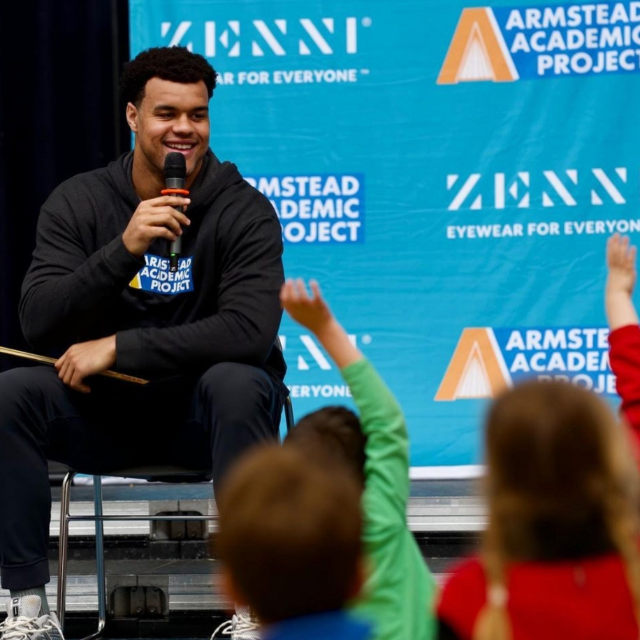 Man holding a microphone in front of a blue &quot;Armstead Academic Project&quot; and &quot;Zenni Eyewear for Everyone&quot; banner.