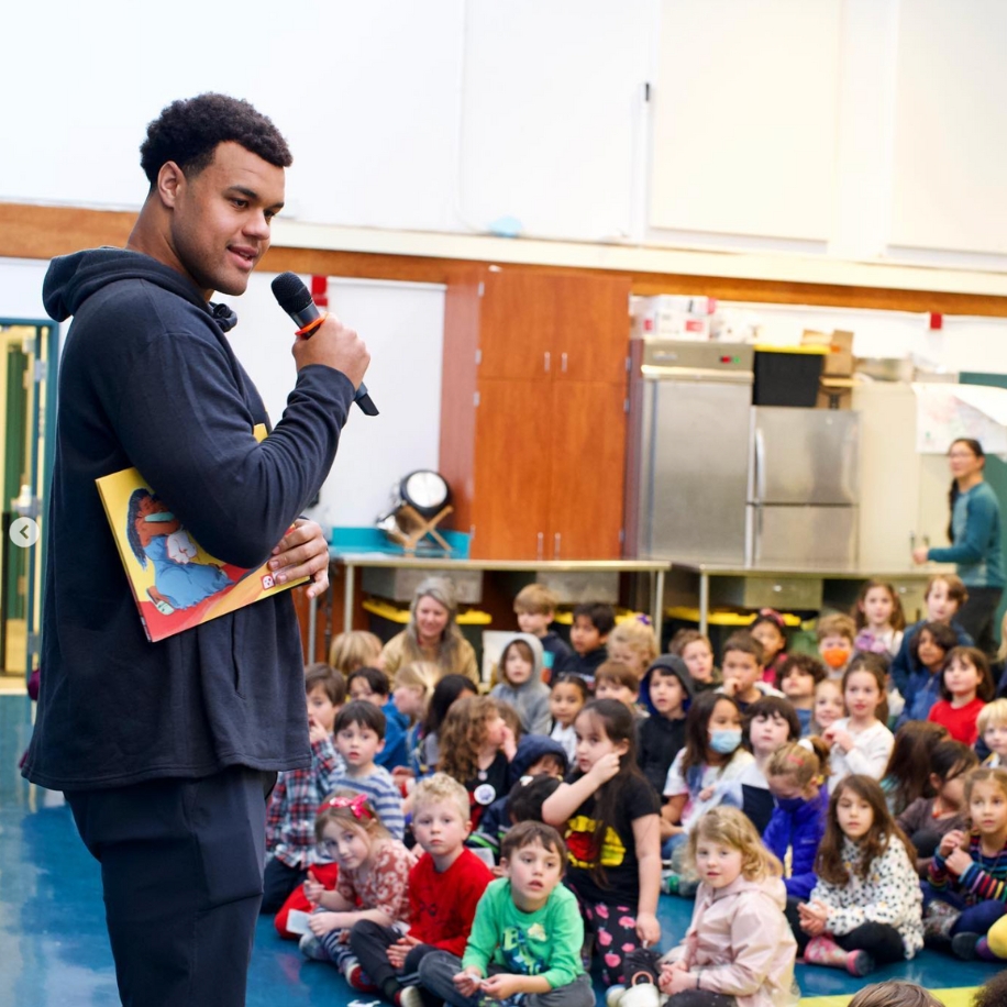 Person holding a book and microphone, speaking to a seated group of attentive children.
