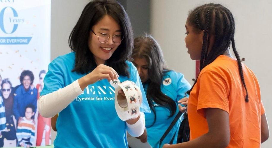 Woman in blue shirt handing roll of sticker glasses to a girl in an orange shirt. Background people blurred.