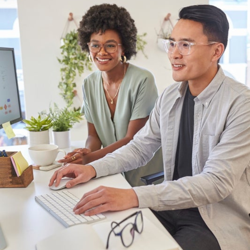 Two people wearing stylish eyeglasses working on a desktop computer in a bright office space.