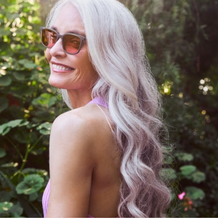Woman wearing dark sunglasses, smiling, with long wavy gray hair and a pink top.