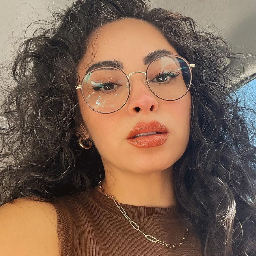 Woman wearing large, round, gold-rimmed eyeglasses, a brown sleeveless top, and a gold chain necklace.