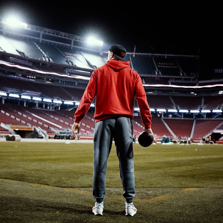 Person in red hoodie and grey pants holding a football on a stadium field at night.