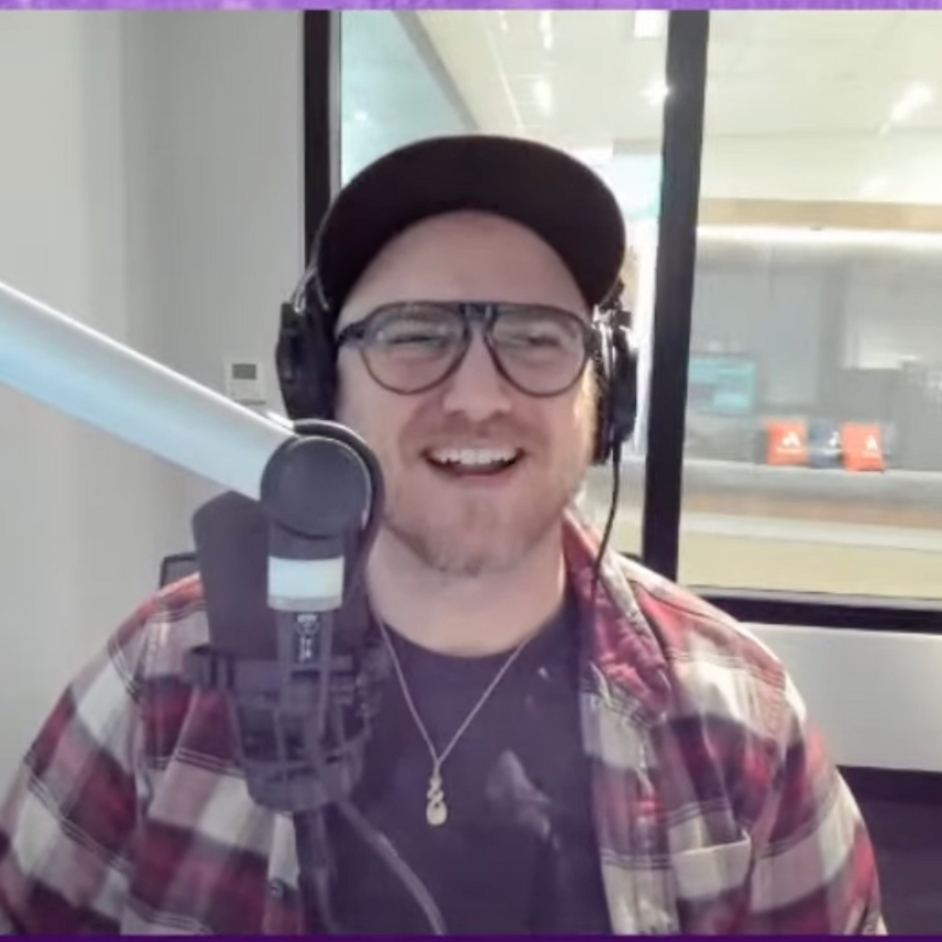 Man wearing glasses and headphones, speaking into a microphone, wearing a checkered shirt and cap.
