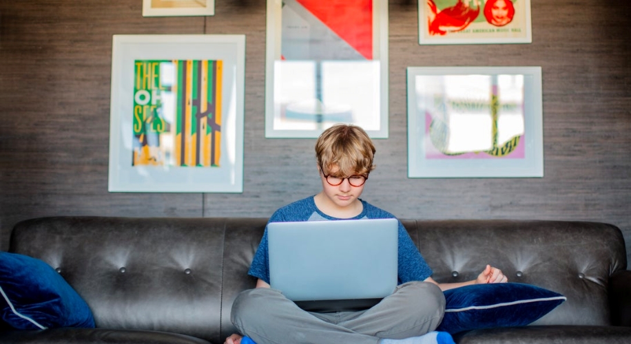 Person using a laptop while sitting cross-legged on a gray couch with colorful artworks in the background.
