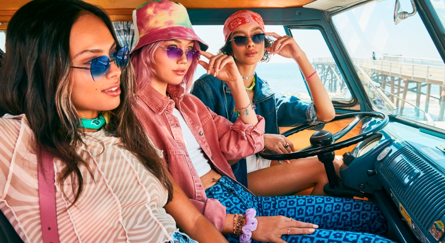 Three people wearing stylish sunglasses and casual outfits, seated inside a vintage blue van.