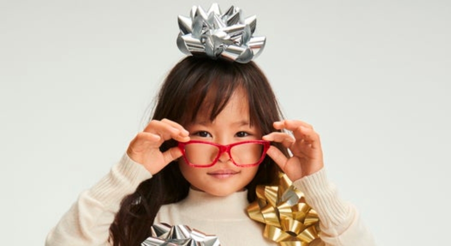 Child wearing red glasses, silver and gold bows, and a cream-colored sweater.