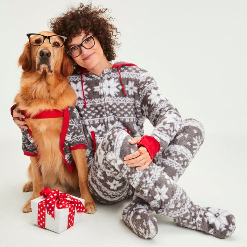 Dog and woman in matching grey snowflake onesies, dog also wears glasses. Present with red ribbon nearby.