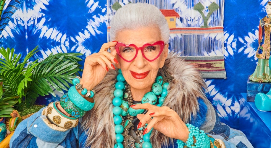 Elderly woman wearing bright heart-shaped glasses, large turquoise beads, various bracelets, and furred outfit.