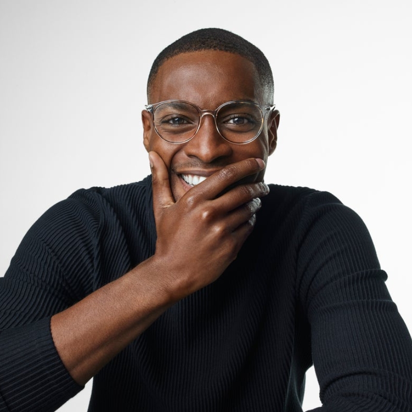 Man wearing clear eyeglasses and a black ribbed sweater, smiling with his hand covering his mouth
