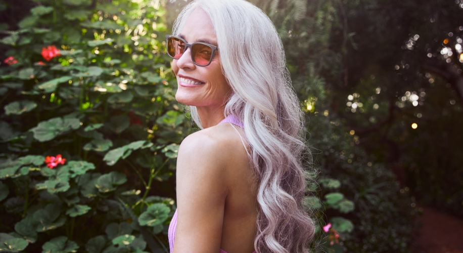 Woman with long silver hair wearing sunglasses and a pink top, smiling outdoors with greenery in the background.