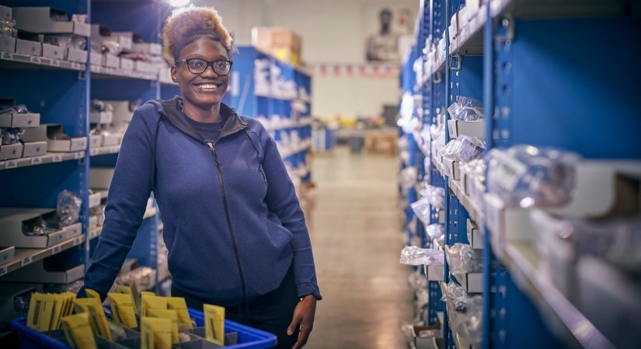 Person standing in an organized warehouse aisle with blue shelves, holding a basket of packaged items.