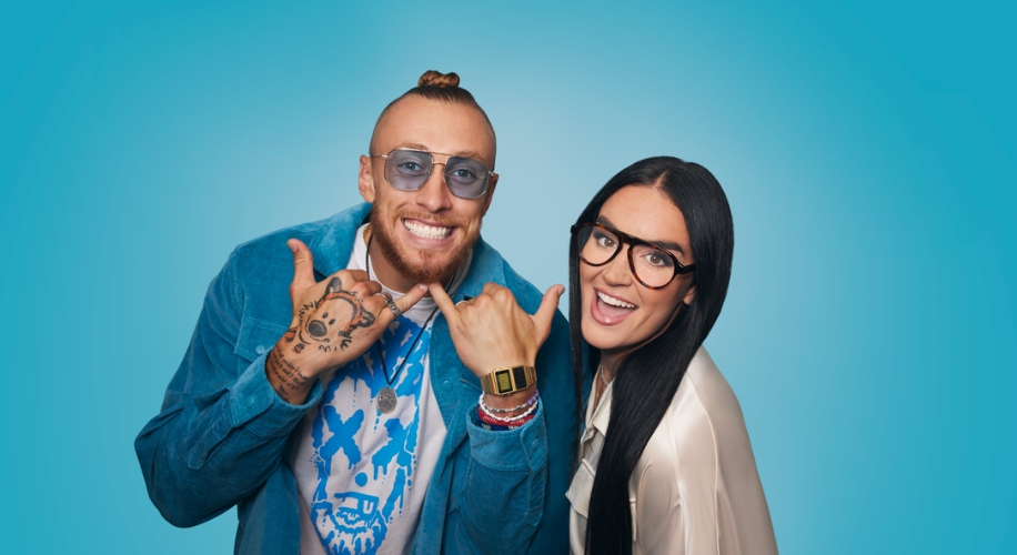 Two people wearing eyewear, a blue shirt, and smiling in front of a blue background.