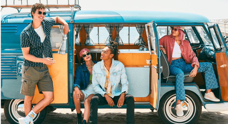 Group of friends relaxing by a vintage blue van. Casual summer outfits with sunglasses and hats.