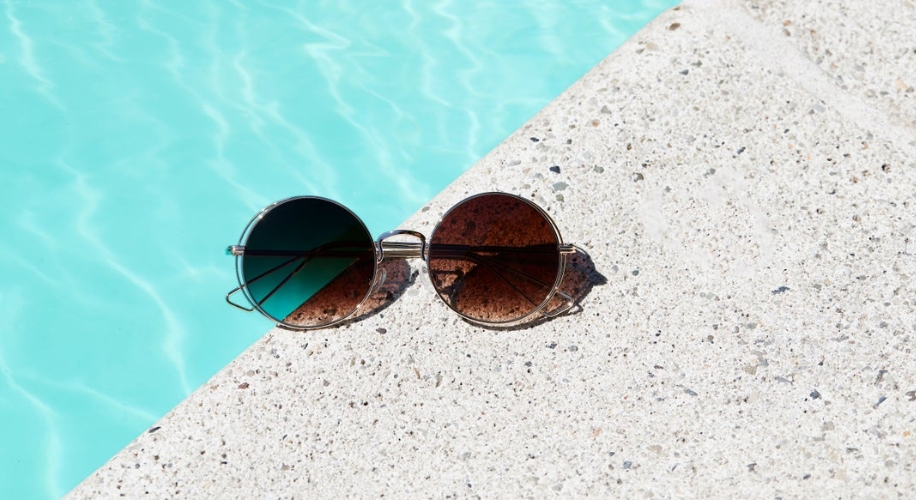 Round sunglasses with dark lenses resting on the edge of a pool with clear blue water.