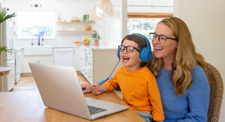 Child wearing blue headphones and an orange shirt, using a laptop with an adult in a blue sweater.