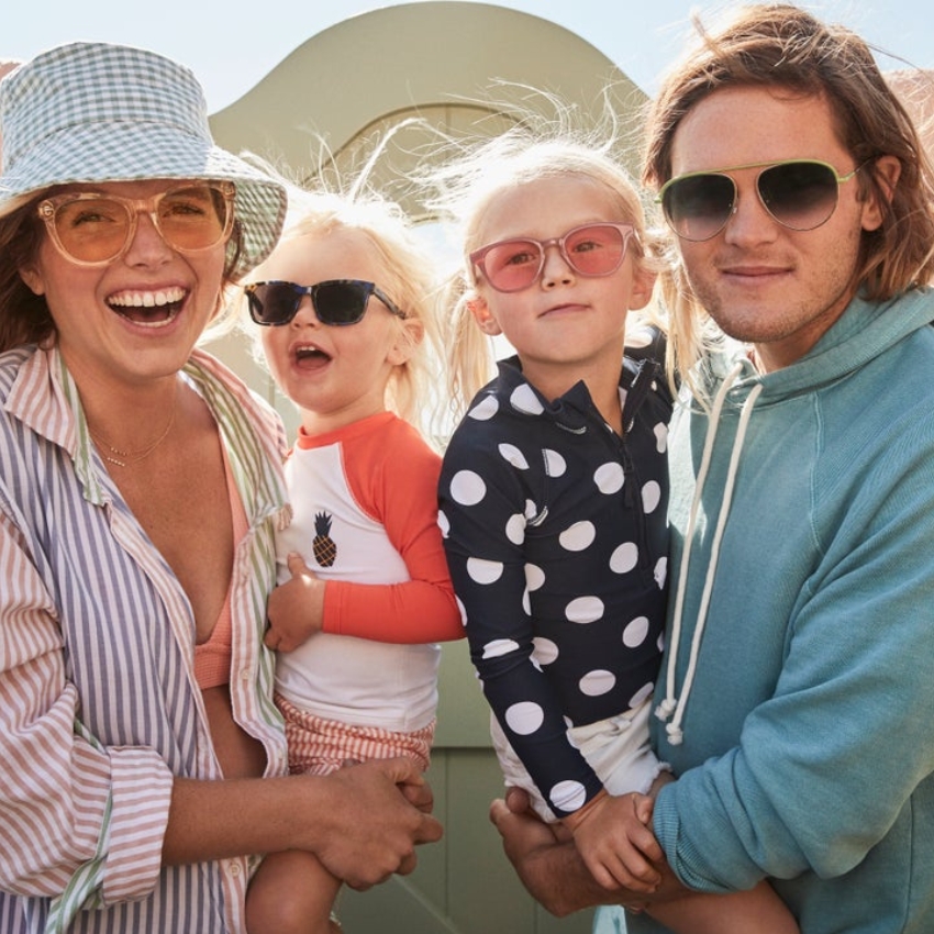 Family wearing sunglasses and beach attire; two adults and two children smiling and posing together.
