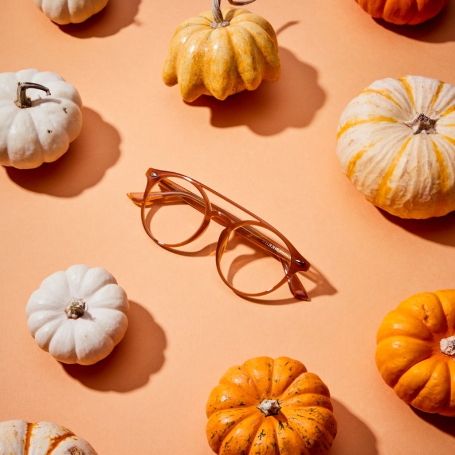 Brown round glasses surrounded by small orange and white pumpkins on a peach-colored background.