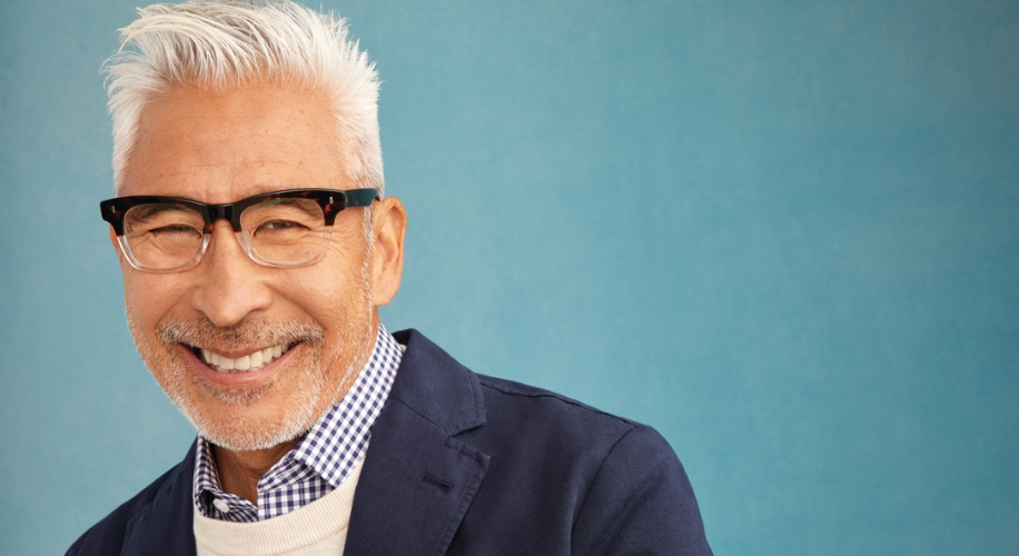 Man wearing glasses, a checkered shirt, a white sweater, and a dark blazer, smiling against a blue background.
