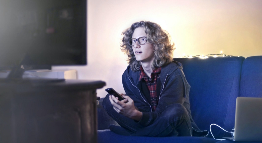 Person with long curly hair and glasses sitting on a blue sofa holding a smartphone, looking at a screen.