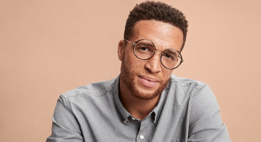 Man wearing round, thin-framed glasses and a light gray button-up shirt against a beige background.