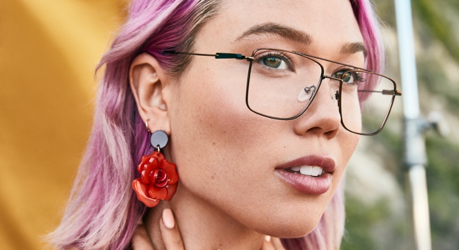 Woman wearing large, square, thin-framed glasses and a red flower earring with purple-tinted hair.