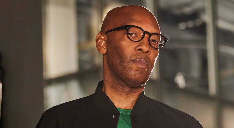 Man wearing black-framed glasses and a black jacket over a green shirt.