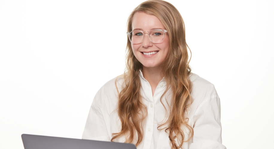 Woman with glasses and long blonde hair smiling, wearing a white shirt, working on a laptop.