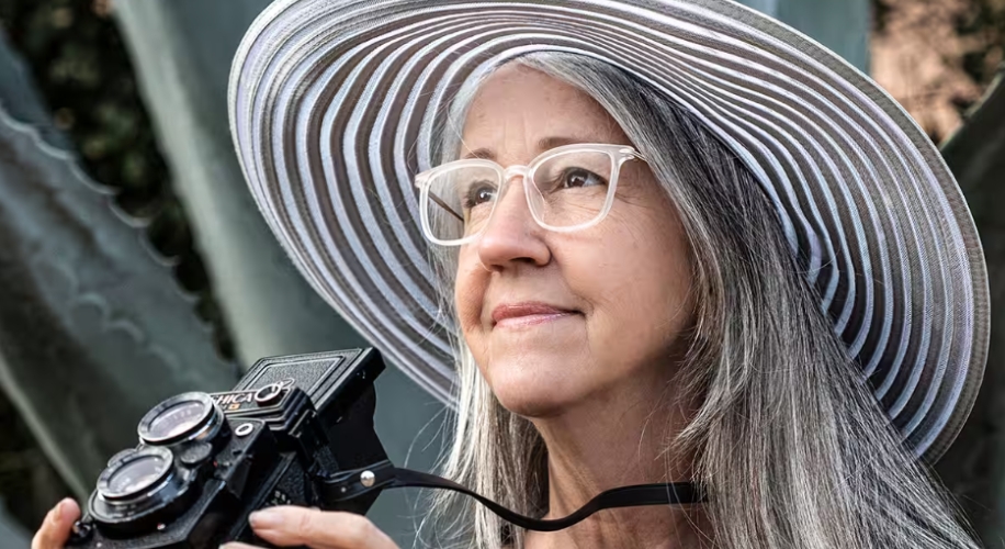 Woman wearing a wide-brimmed, striped hat and glasses, holding a vintage Yashica-Mat camera.