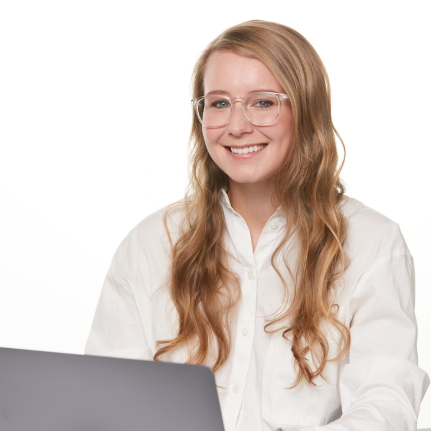 Woman with long wavy hair wearing clear glasses and a white shirt, smiling while using a laptop.