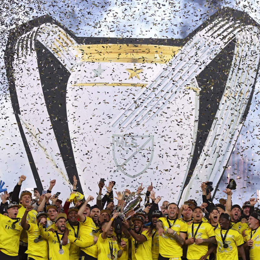 Soccer team in yellow uniforms celebrating with an MLS trophy amid a shower of confetti.