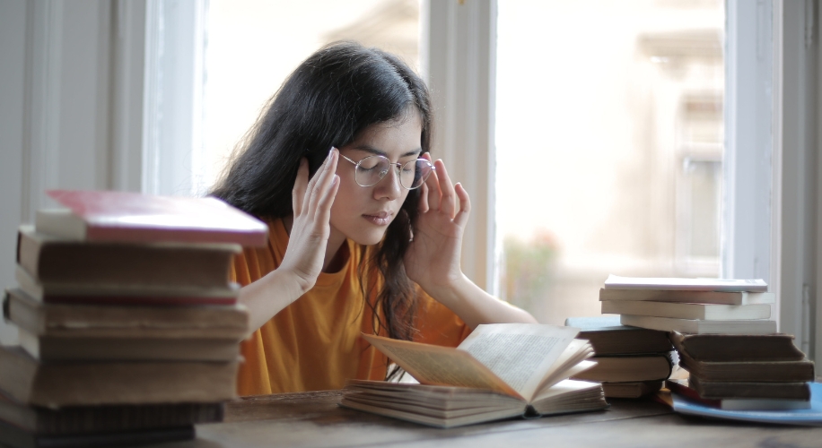 Woman with glasses reading a book, surrounded by stacks of books.