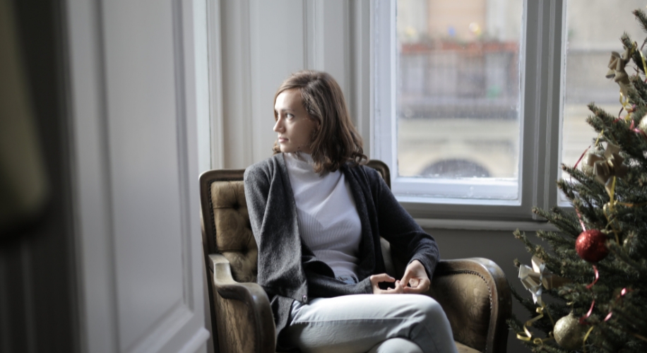 Person sitting on a vintage armchair near a window, gazing outside, with a decorated Christmas tree nearby.