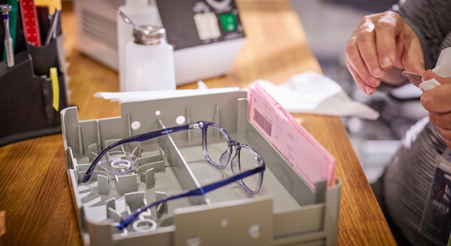 Blue eyeglasses in a gray tray on a wooden table next to a person&#039;s hands and pink documents.