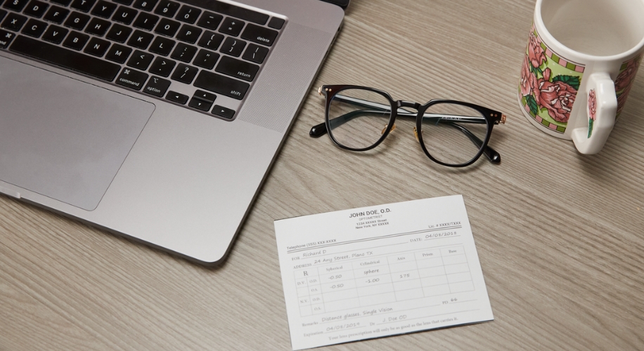 Prescription glasses with black frames, eyeglass prescription from JOHN DOE, O.D., laptop keyboard, floral mug.
