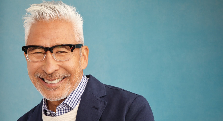 Man wearing black-rimmed glasses, a checkered shirt, and a dark blazer against a blue background, smiling.