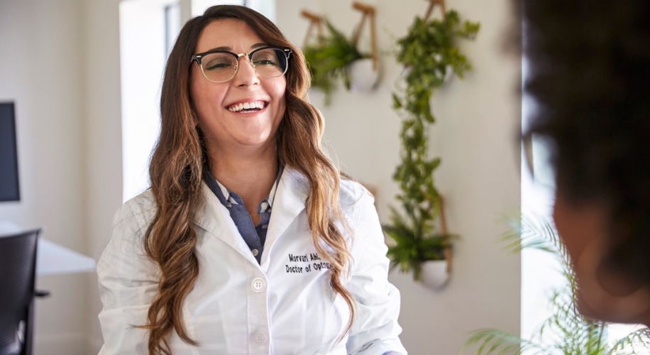 Doctor of Optometry in a white coat, smiling and wearing glasses. Text on coat: &quot;Marvez Ahn, Doctor of Optometry.&quot;
