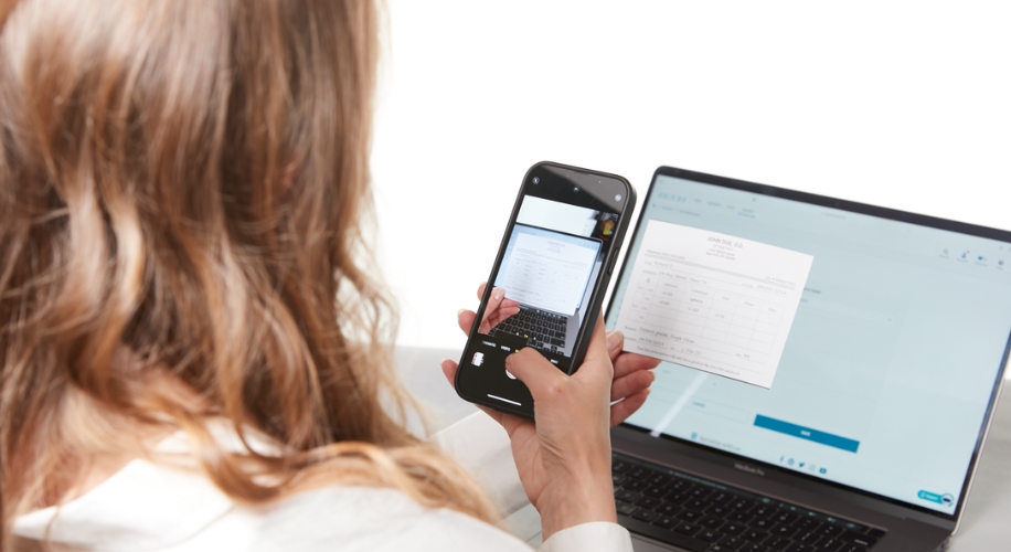 Person holding a phone, capturing a document displayed on a laptop screen while holding the document in hand.