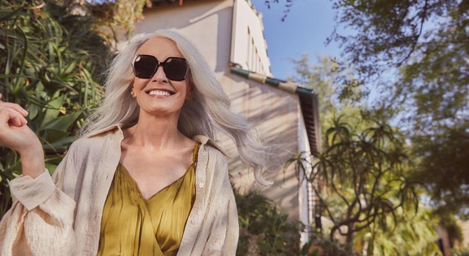 Woman wearing black sunglasses, beige shirt, and olive green dress smiles outdoors.