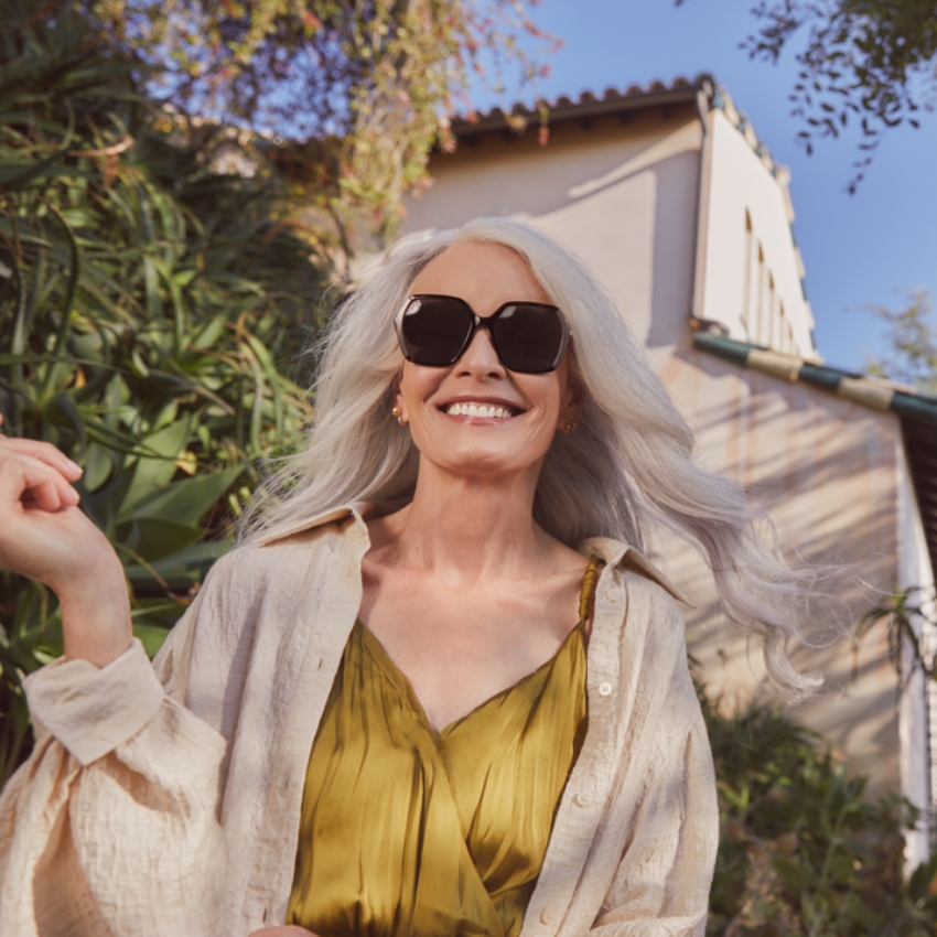 Woman wearing oversized black sunglasses and a light beige open buttoned shirt over a green dress.