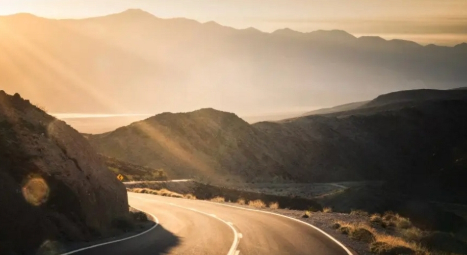Sunlit winding road through mountainous terrain with distant hills under a clear sky.